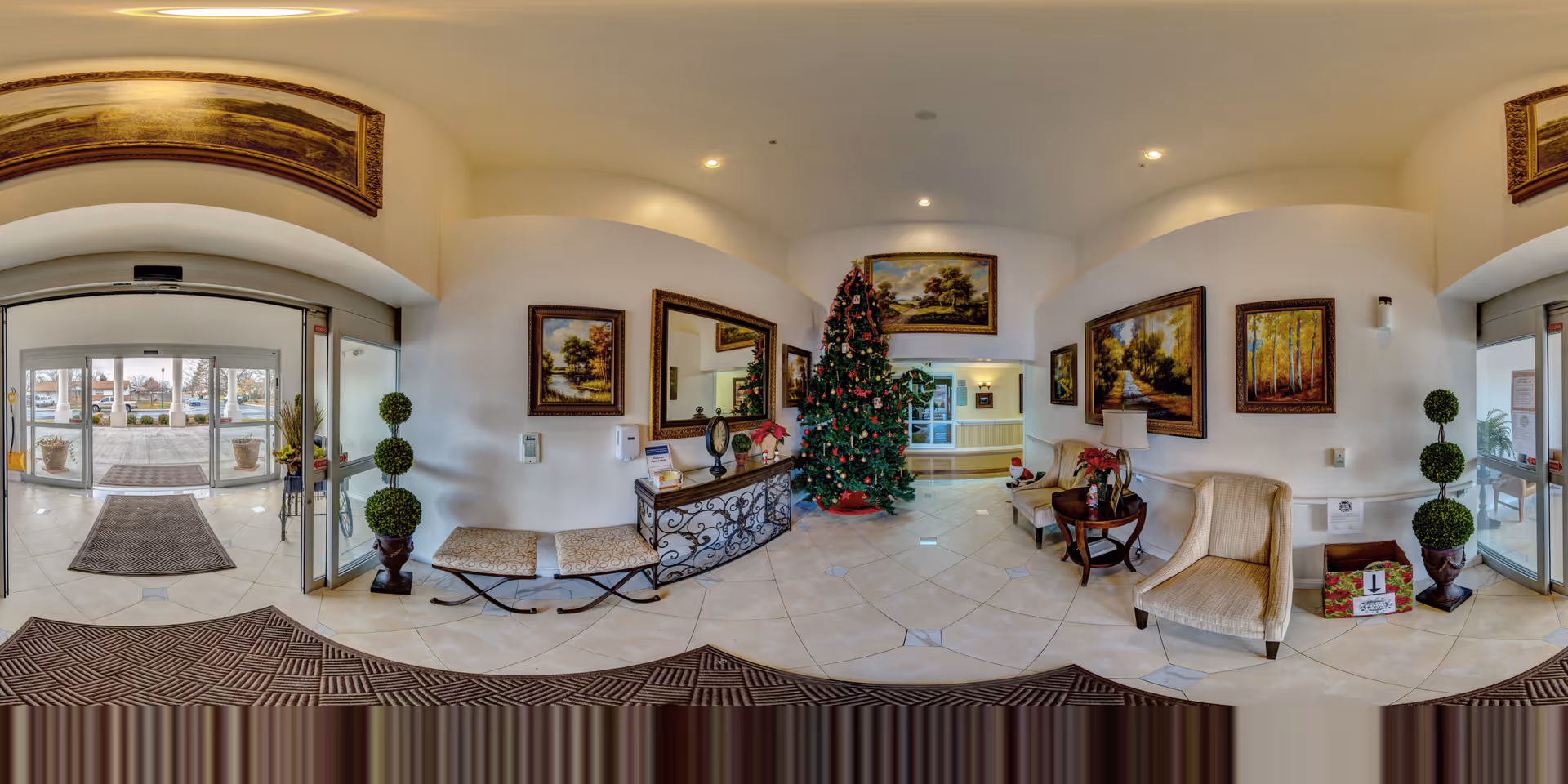 A spacious and well-lit lobby area decorated for Christmas with a large Christmas tree in the center. The room features tiled floors, several framed landscape paintings on the walls, a decorative console table with a mirror above it, cushioned benches, and comfortable armchairs. There are potted plants on either side of the entrance doors, and gift boxes placed near one of the chairs. The entrance has glass sliding doors leading outside.