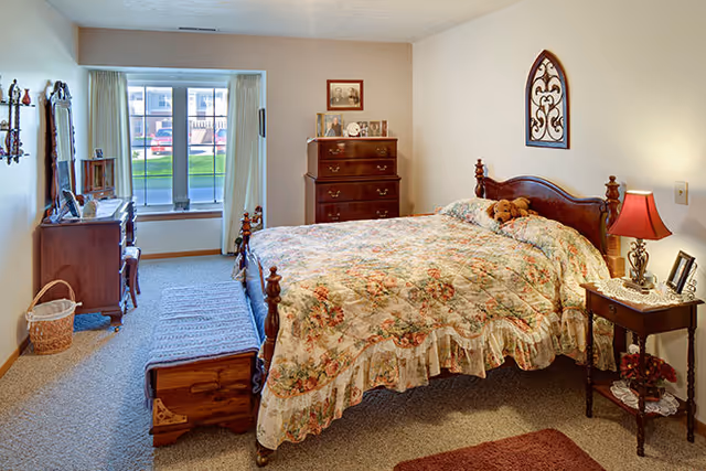 A cozy bedroom with a floral-patterned bedspread on a wooden bed frame. There is a wooden chest at the foot of the bed, a nightstand with a red lamp and framed photos, a dresser with decorative items, and a window with curtains letting in natural light. The walls are light-colored and there is a carpeted floor.
