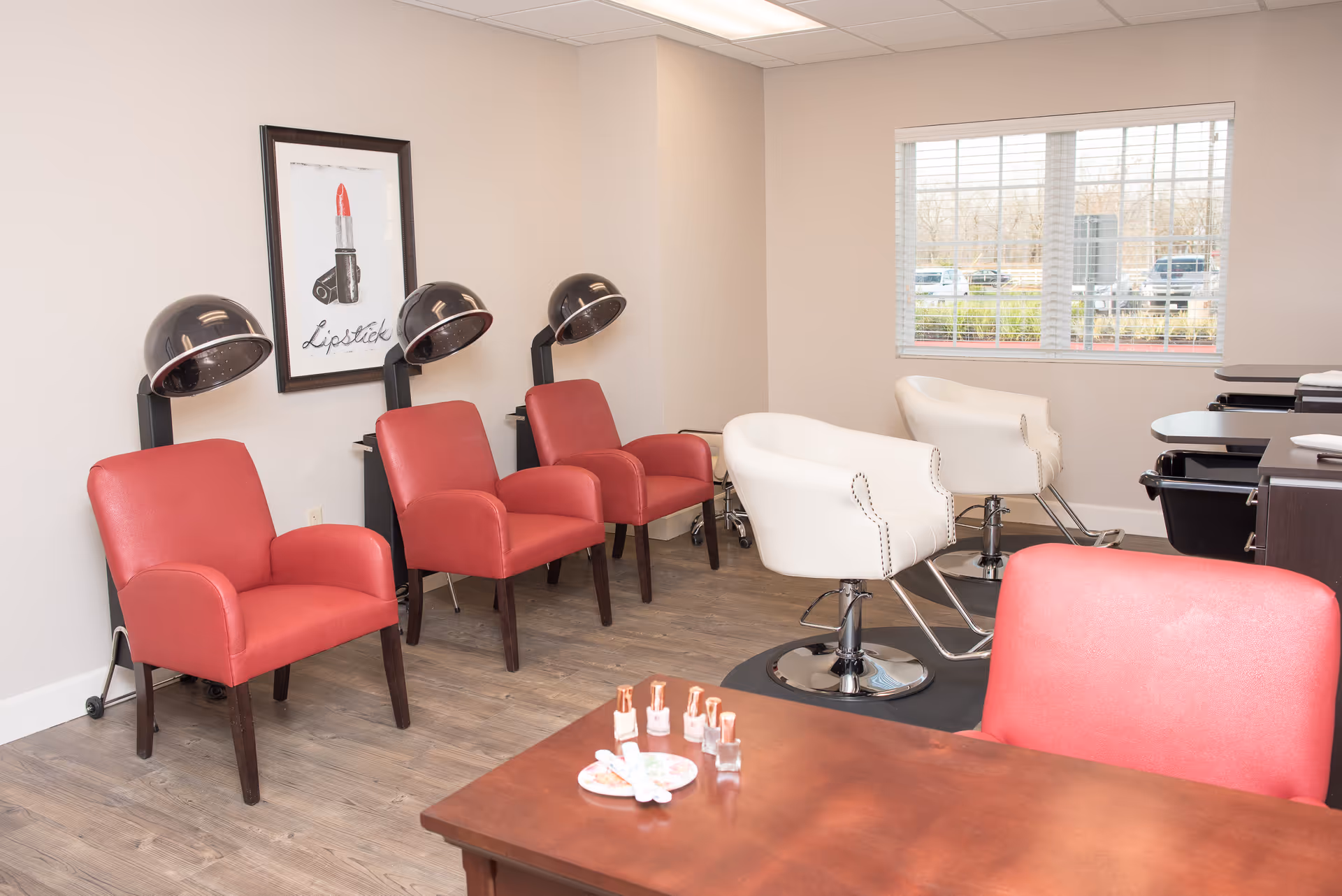 A bright salon room with red waiting chairs, white styling chairs under hooded dryers, a manicure table with nail polish, and a window.