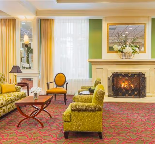 A cozy living room area with a lit fireplace, green upholstered armchairs, a patterned sofa, a wooden coffee table with a small flower arrangement, and a large window with sheer white curtains and yellow drapes. A mirror and a floral arrangement are placed above the fireplace.