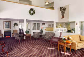 A spacious assisted living facility common area with patterned carpet, multiple seating arrangements including floral upholstered chairs and sofas, wooden tables with flower vases, framed artwork on the walls, a fireplace, and a decorative wreath hanging on the upper wall.