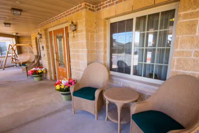 Covered front porch with wicker chairs and a small table, potted flowers, and a wooden double-door entrance.