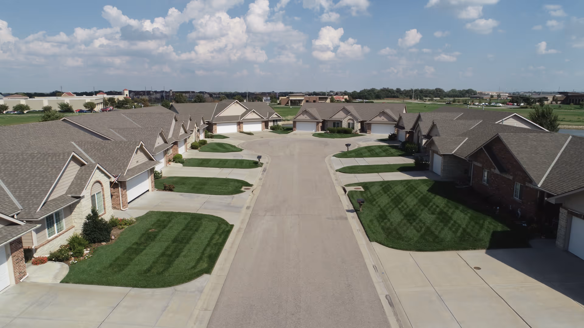 Aerial view of a residential street in The Villas at Hampton Lakes featuring single-story homes with garages, neatly trimmed lawns, and a clear blue sky with scattered clouds.