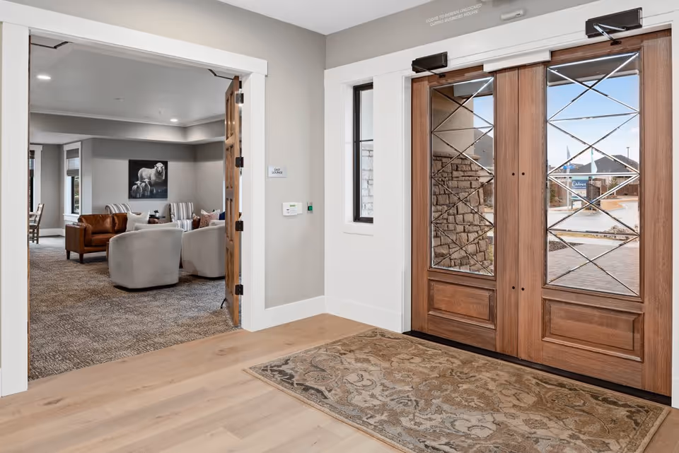 Entrance area with wooden double doors featuring glass panels with a crisscross design. Inside, there is a carpeted lounge area with several armchairs and a painting of sheep on the wall. The floor near the entrance is light wood with a patterned rug.