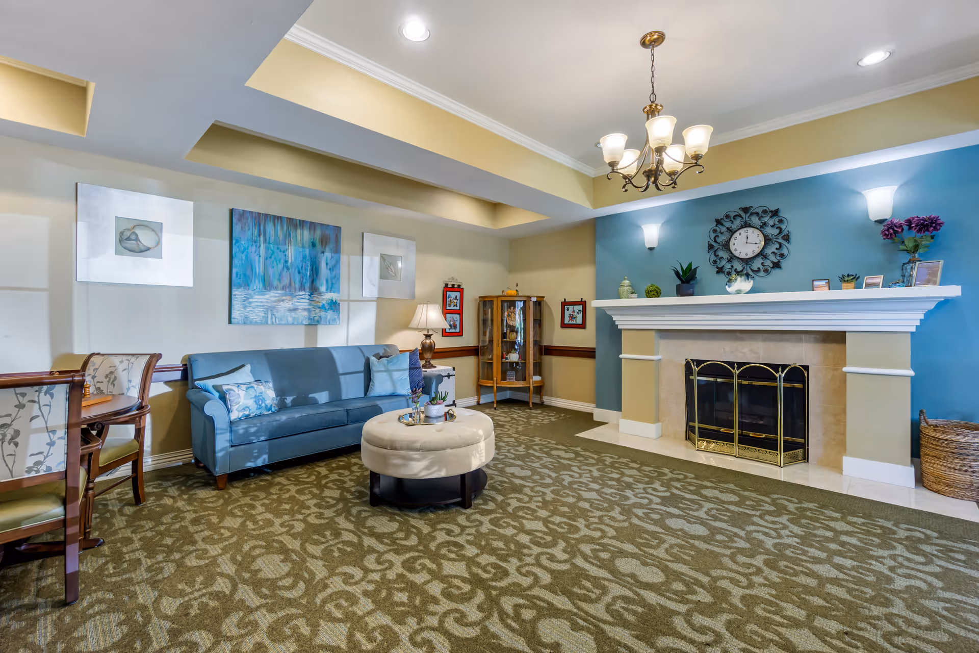 A cozy living room area with a blue couch adorned with pillows, a round beige ottoman in the center, and a fireplace with a decorative clock and plants on the mantel. The walls are painted in soft yellow and blue tones, and there is a chandelier hanging from the ceiling. A small wooden dining table with chairs is visible on the left side, along with framed artwork on the walls.