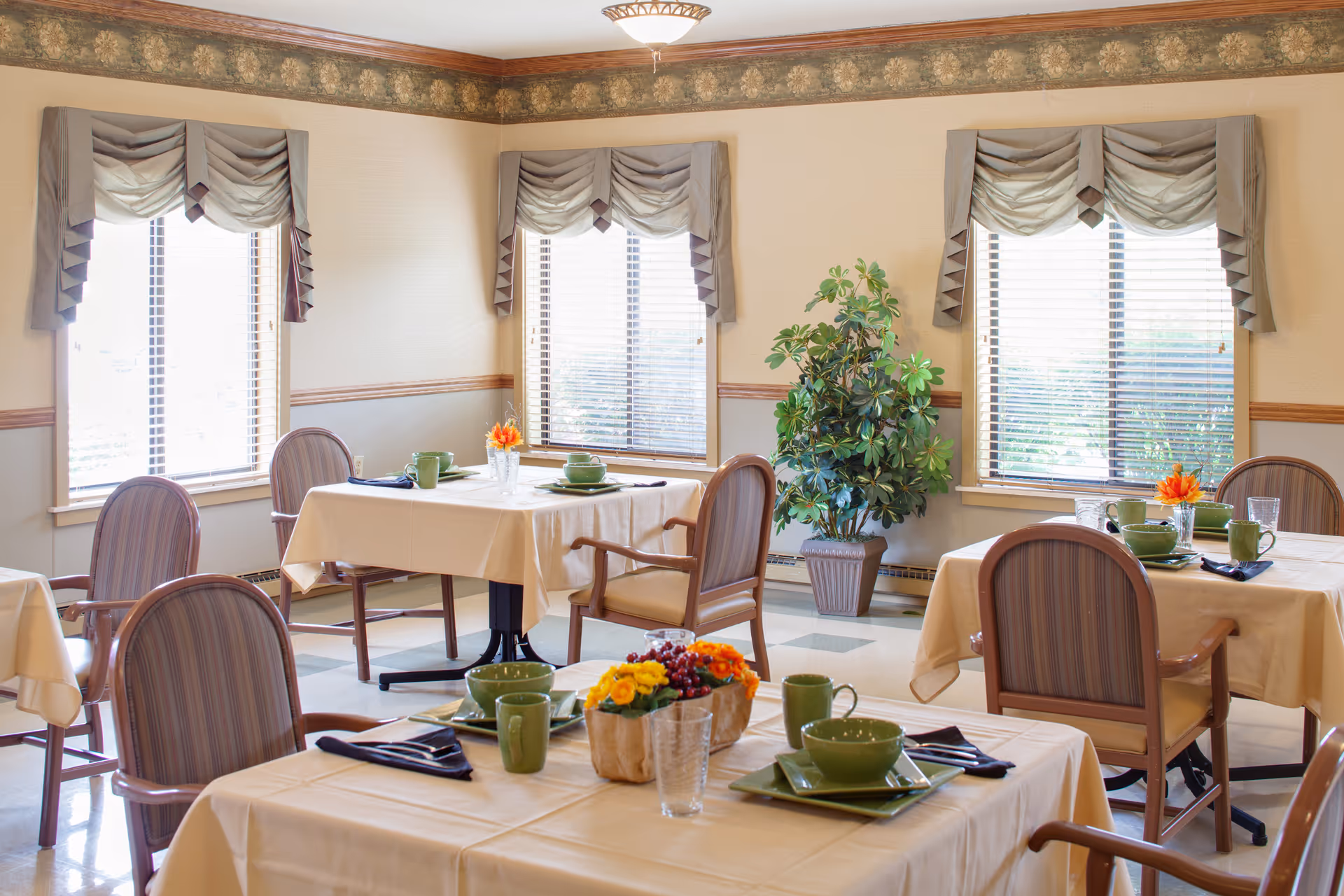 Bright dining room with multiple tables set with green dishware, chairs, large windows with valances, and a potted plant.