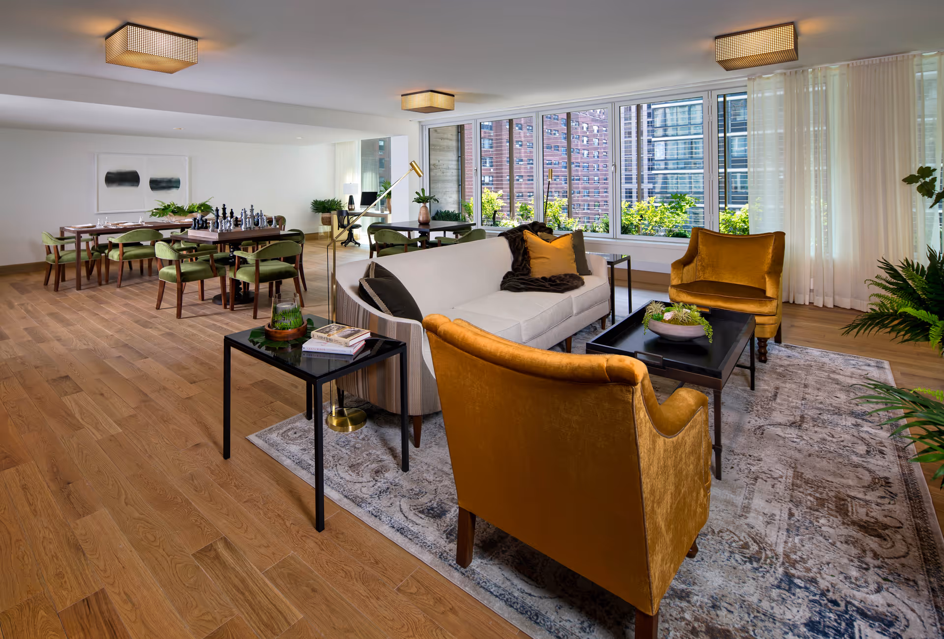 A spacious and modern living room area with large windows showing a cityscape outside. The room features a white sofa with decorative pillows, two mustard yellow armchairs, a black coffee table with a plant centerpiece, and a side table with books and a glass terrarium. In the background, there is a dining area with a wooden table and green upholstered chairs, a chess set on the table, and contemporary ceiling lights. The floor is wooden with a patterned rug under the seating area, and there are several plants adding greenery to the space.