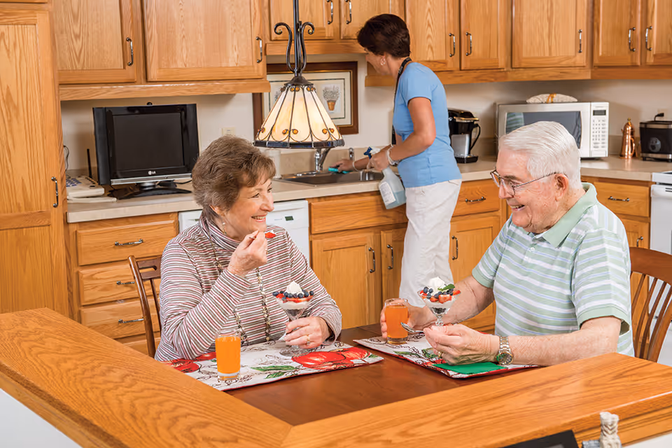 An elderly man and woman sit at a wooden kitchen table enjoying dessert parfaits and orange drinks, smiling at each other. In the background, a woman in a blue shirt and white pants is cleaning the kitchen sink. The kitchen has wooden cabinets, a microwave, and a small television on the counter.