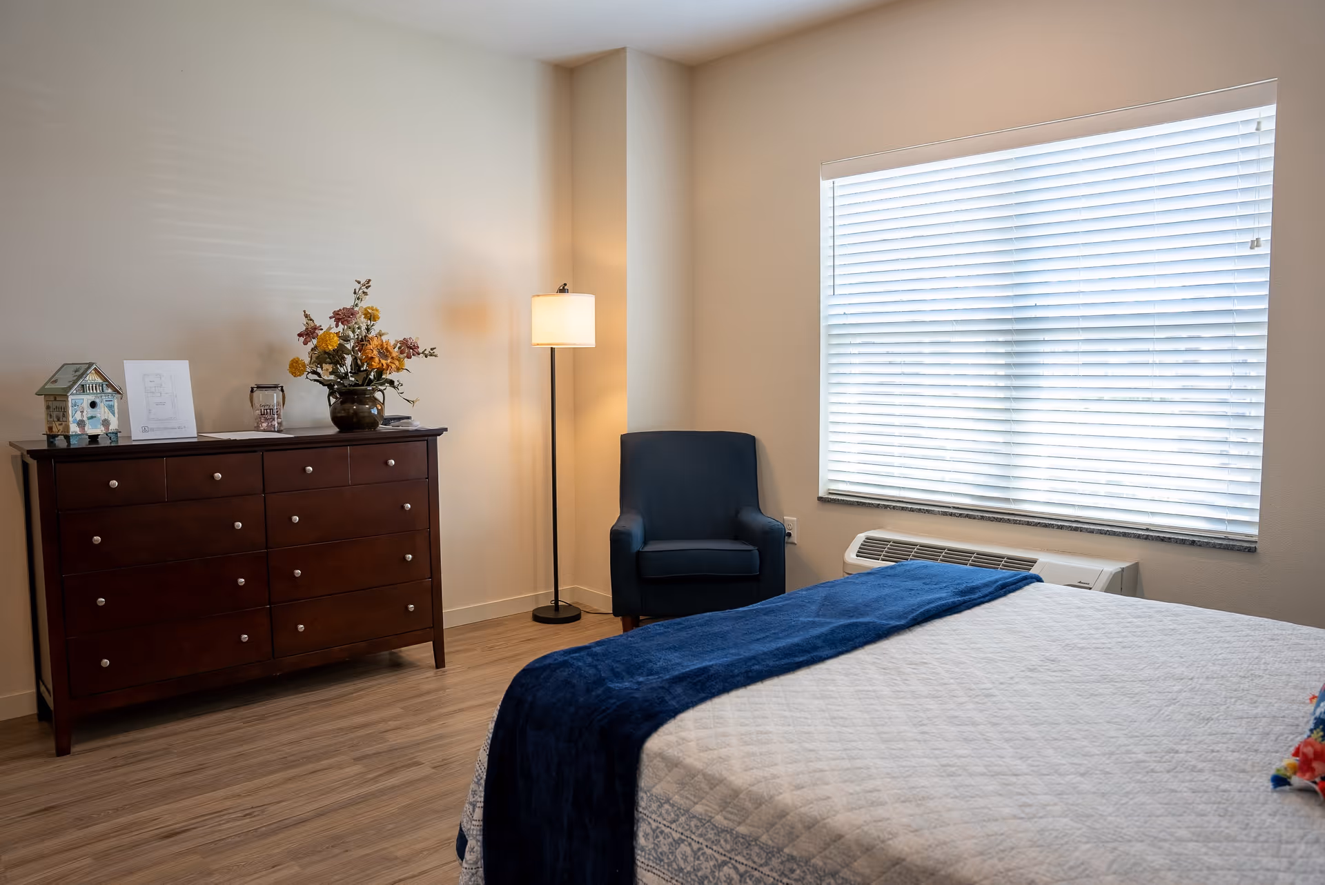 Well-lit bedroom with a bed, blue armchair, wooden dresser with flowers, floor lamp, and a large window with blinds.