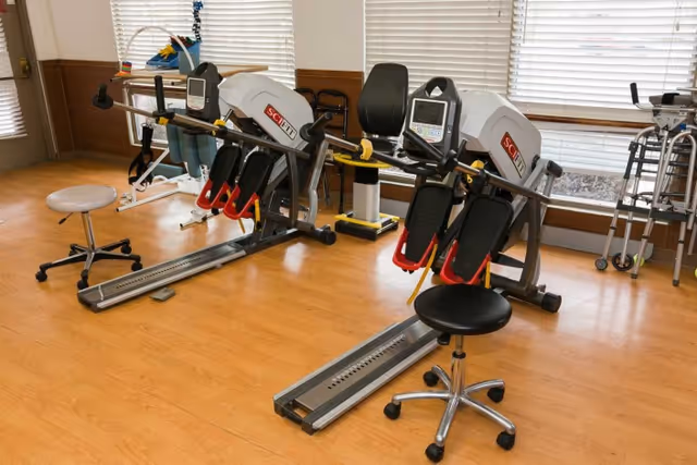 A rehabilitation room with exercise equipment including two SciFit machines, a rolling stool, and walkers near windows with blinds. The room has wooden flooring and light-colored walls with wooden paneling.