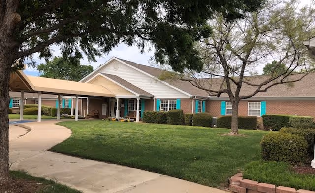 Front exterior of a single-story brick senior living facility with a covered entrance, green lawn, trees, and turquoise window shutters.