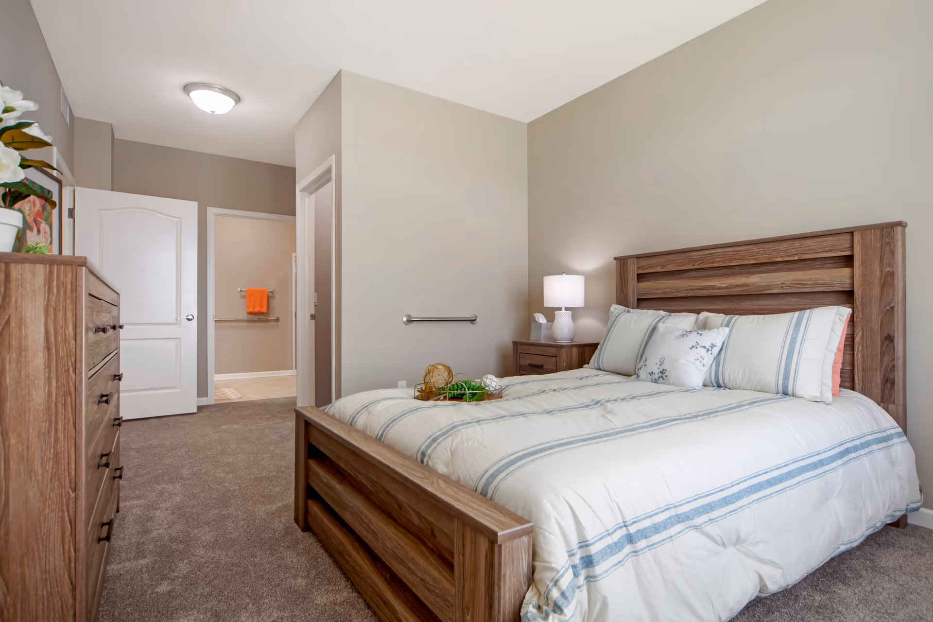 Bright furnished bedroom featuring a wooden bed with striped bedding, a nightstand with lamp, dresser, and an open door to a hallway.