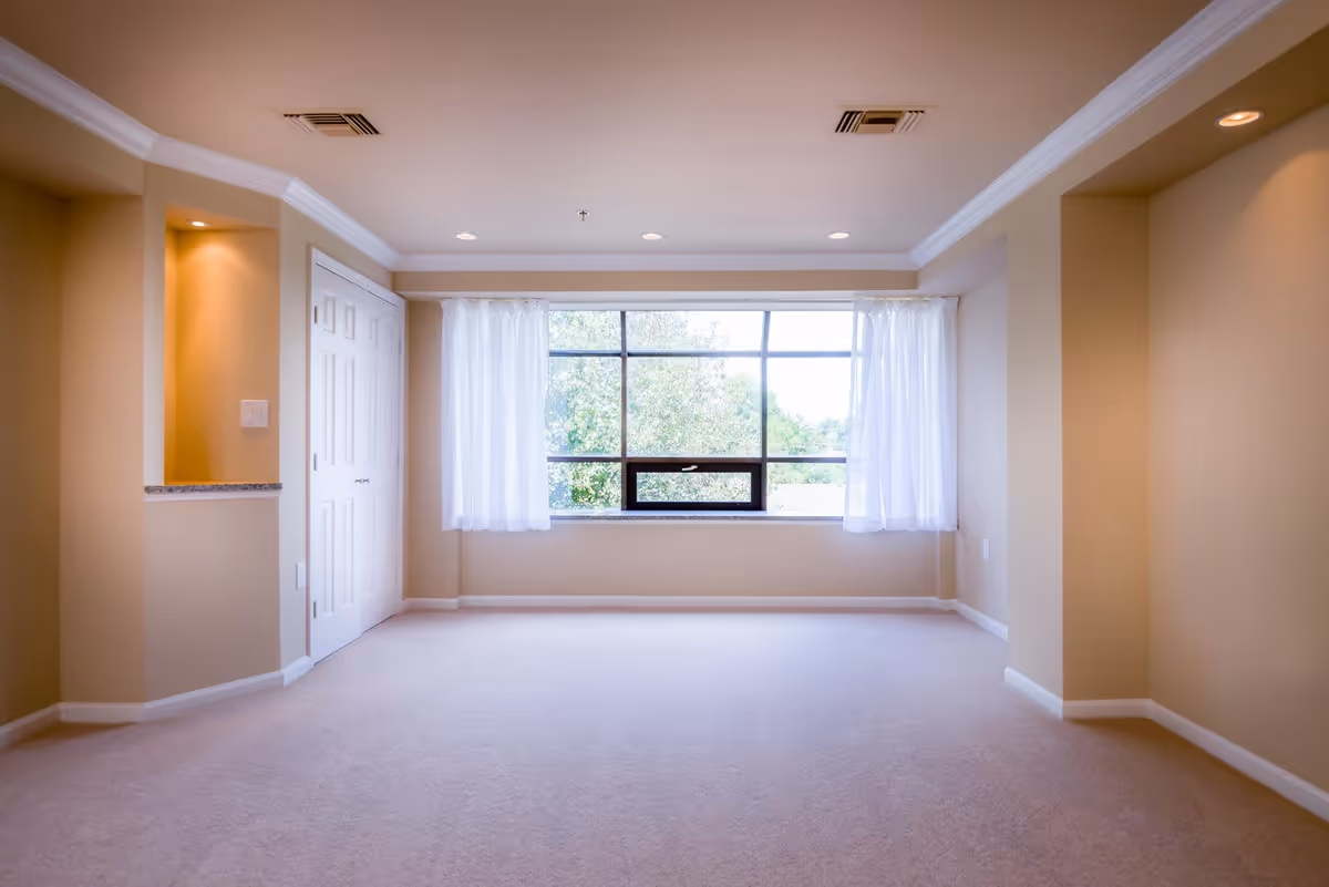 Empty room with beige walls and carpet, a large window with white curtains letting in natural light, recessed ceiling lights, and a white double-door closet on the left side.