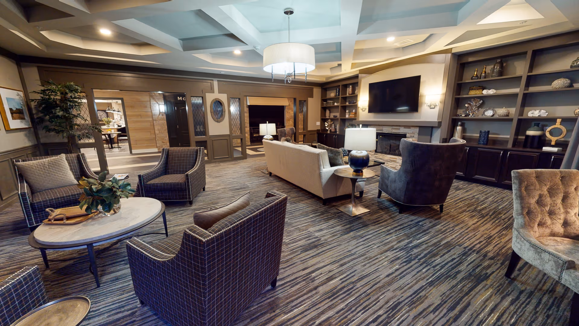 A spacious and elegantly furnished living room area in a senior living facility. The room features a large beige sofa, several armchairs with different upholstery patterns, a round coffee table with a plant centerpiece, built-in shelves with decorative items, a flat-screen TV mounted above a fireplace, and soft lighting from table lamps and a ceiling fixture. The room has a patterned carpet and a coffered ceiling design.