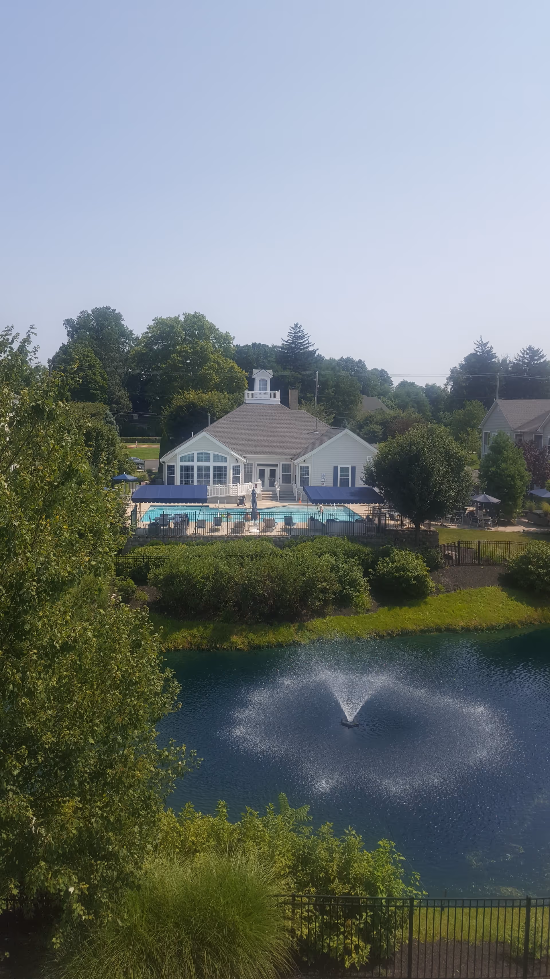 View of a senior living facility building with a swimming pool in front, surrounded by a fence and shaded seating areas. In the foreground, there is a pond with a water fountain spraying water in a circular pattern, and lush green trees and bushes around the pond.