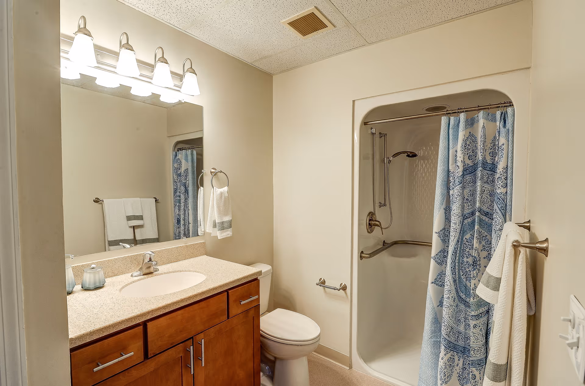 A clean bathroom featuring a wooden vanity with a beige countertop and an oval sink. Above the sink is a large mirror with four light fixtures. To the right is a white toilet and a shower with a blue and white patterned curtain. The walls are light-colored, and there are towel racks holding white towels with green stripes.