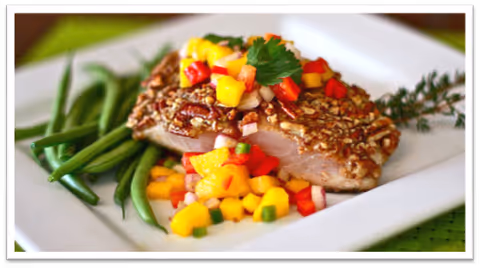 A plated meal featuring a pecan-crusted piece of fish topped with a colorful mango salsa, served alongside green beans on a white square plate with a green placemat underneath.
