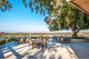 Outdoor patio area with multiple tables and chairs arranged on a concrete surface under a large tree, overlooking a scenic view of distant hills and clear blue sky.