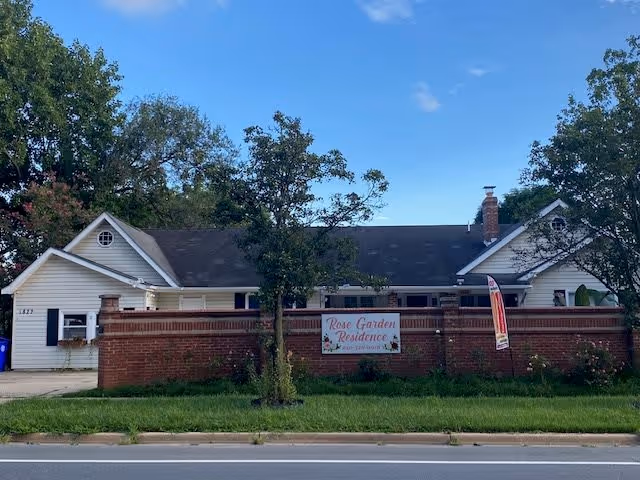 Exterior view of a single-story residential building with white siding and a dark roof behind a low red brick wall. A sign on the wall reads 'Rose Garden Residence'. There are trees and grass in front of the building, and a clear blue sky above.