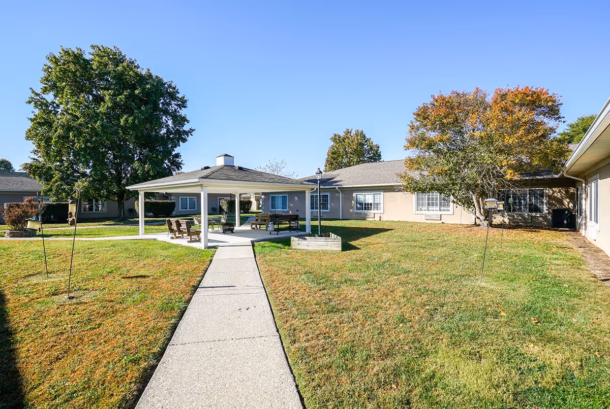 Outdoor courtyard area at Rolling Meadows Place featuring a paved walkway leading to a covered seating area with chairs and a picnic table. Surrounding the courtyard are single-story buildings with windows, green grass, and several trees with some autumn foliage under a clear blue sky.