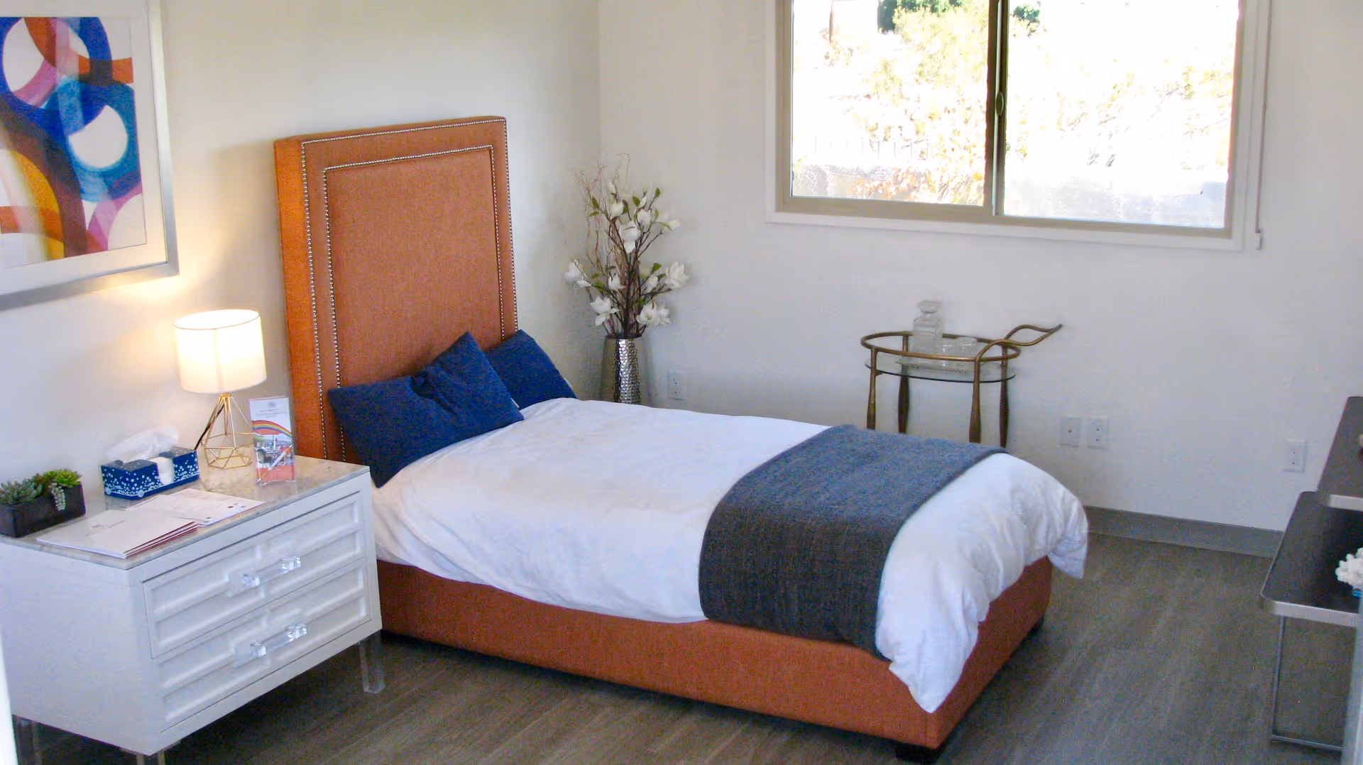 A neatly made single bed with an orange upholstered headboard and blue pillows in a bright room. Next to the bed is a white nightstand with a lamp, tissue box, and some papers. A colorful abstract painting hangs on the wall above the nightstand. There is a window letting in natural light and a small round table with decorative items near the window. The floor is wood, and the walls are white.