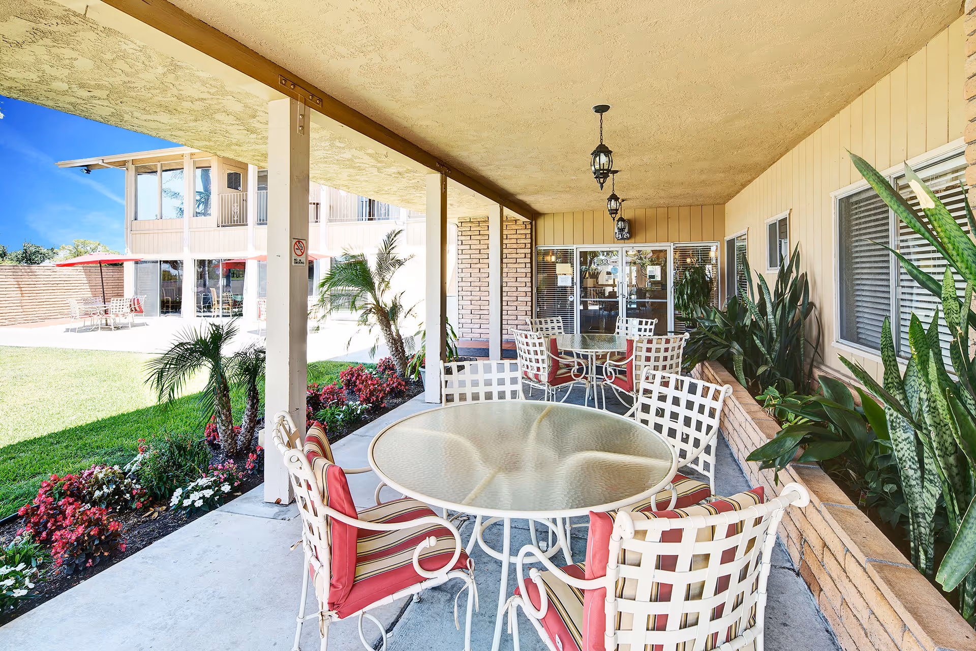 Covered outdoor patio area with round glass tables and white metal chairs with red and beige striped cushions. The patio is adjacent to a building with windows and plants along the wall. In the background, there is a grassy lawn with flower beds, palm trees, and another building with large windows and red umbrellas over tables.