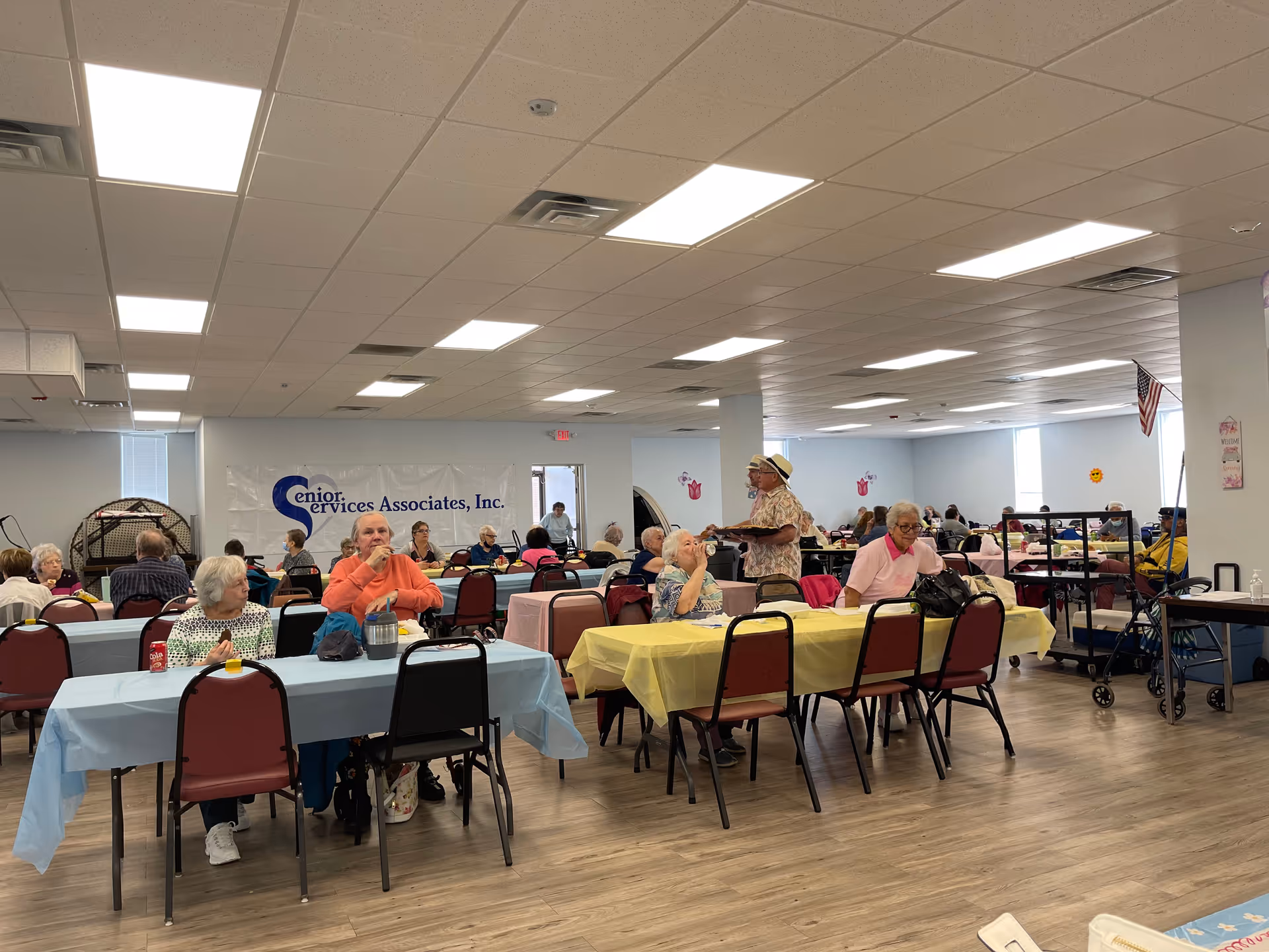 A large dining room in an assisted living facility with several elderly residents seated at tables covered with pastel-colored tablecloths. Some residents are eating or drinking, while others are conversing. The room has a light-colored wood floor, white ceiling with fluorescent lights, and light blue walls. A banner on the wall reads 'Senior Services Associates, Inc.' and an American flag is visible in the background.