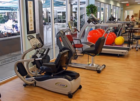 Interior view of a fitness room in a senior living facility with exercise equipment including a recumbent bike, treadmill, and large exercise balls. The room has large windows letting in natural light and a wooden floor. There are chairs and plants visible, and people can be seen in the background near the entrance.