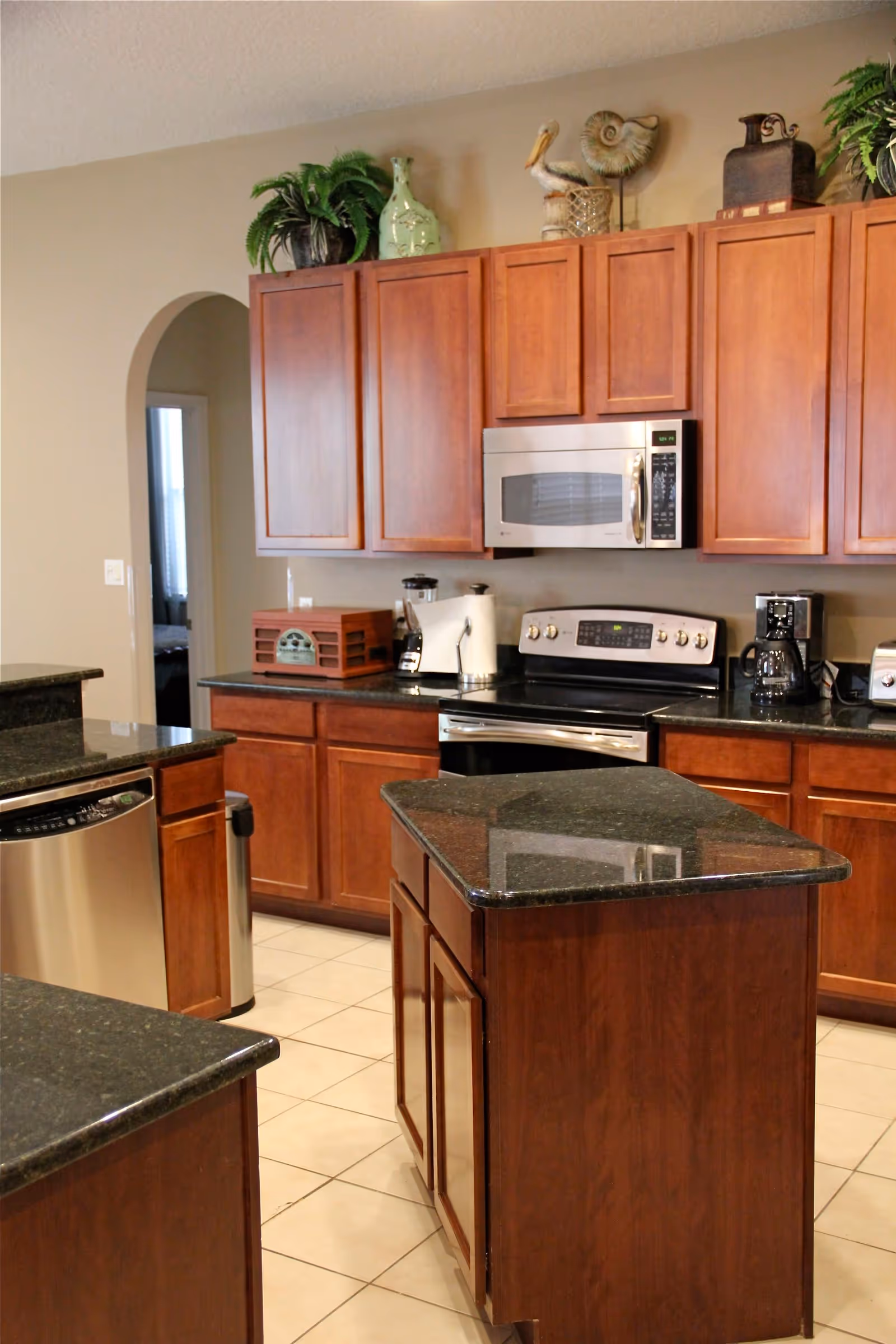 A kitchen with wooden cabinets, a black granite countertop island, stainless steel appliances including a microwave, stove, dishwasher, and coffee maker. There are decorative items and plants on top of the cabinets, and a tiled floor.