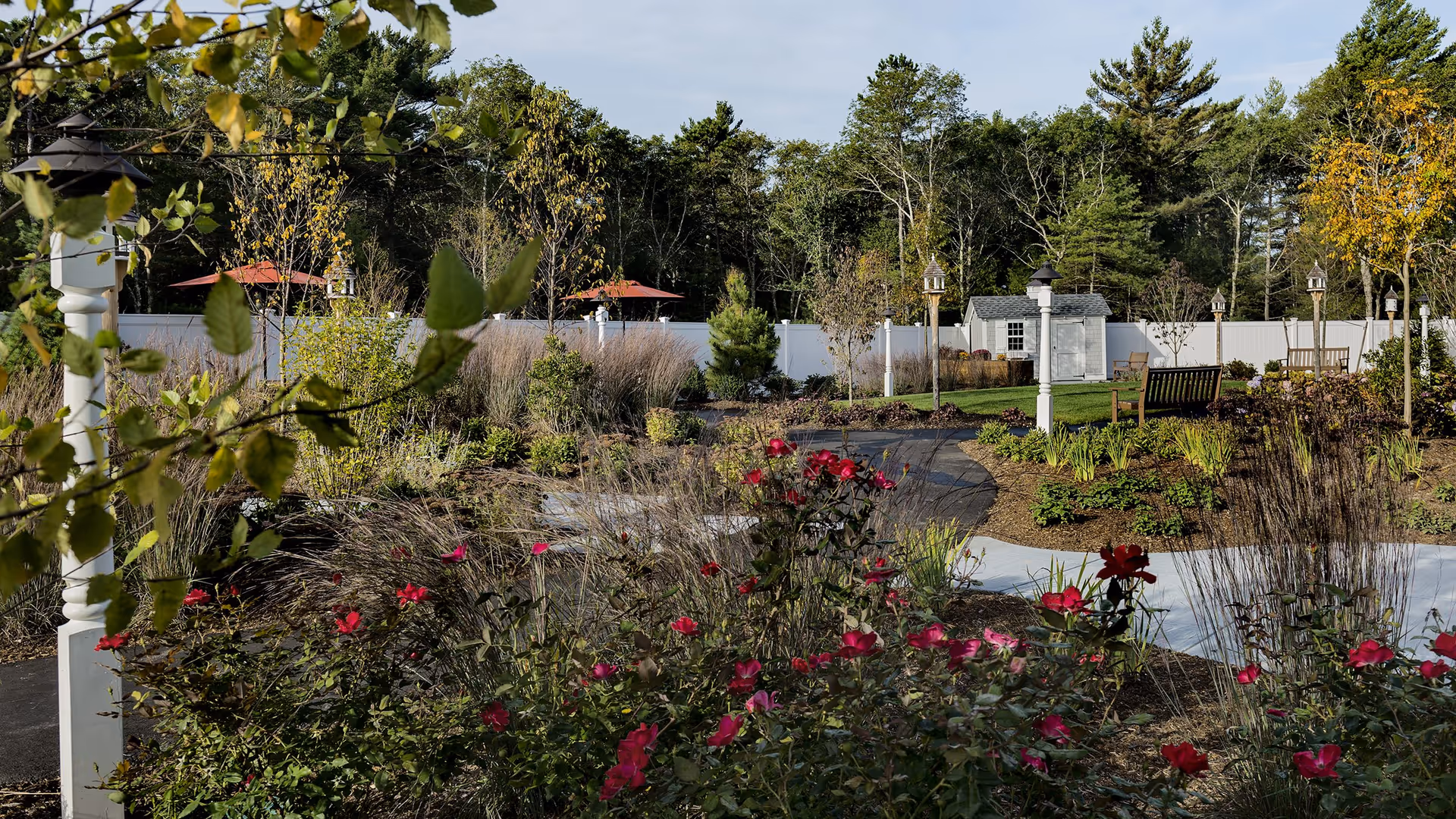 A landscaped outdoor garden area with a winding paved pathway, red flowers, various plants, trees, benches, lamp posts, and a small white shed in the background, surrounded by a white fence and tall trees.