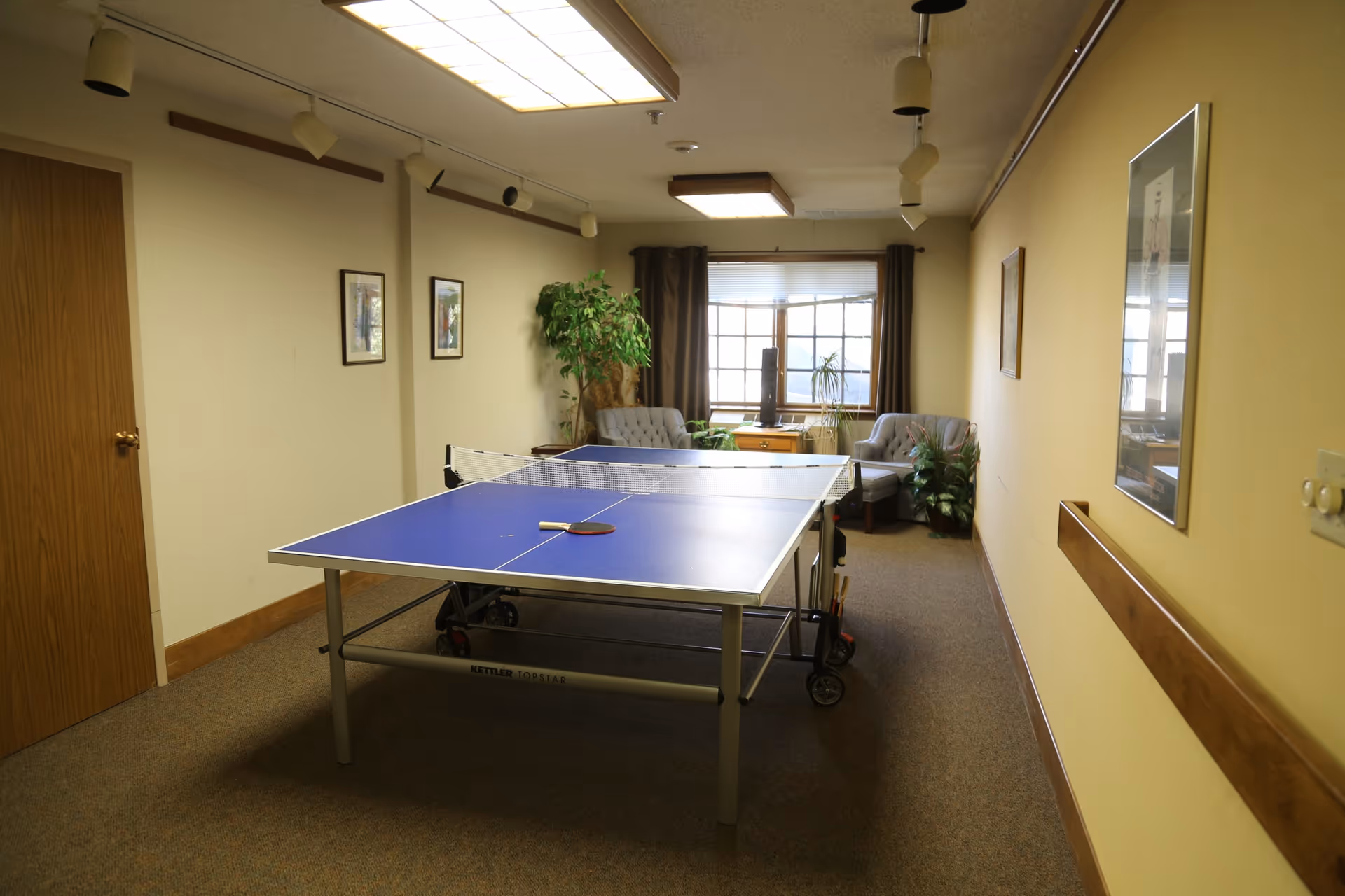 Indoor room with a blue ping pong table in the center, two gray armchairs near a window with brown curtains, several framed pictures on the walls, and some potted plants. The room has beige walls and carpeted floor with overhead fluorescent lighting.