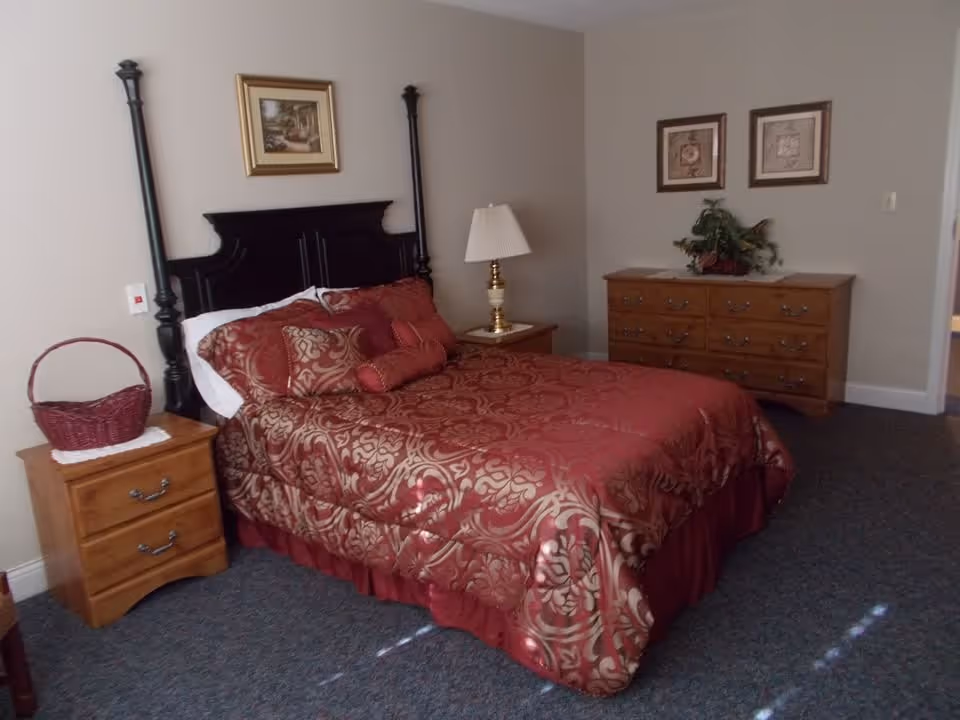 A bedroom with a large bed covered in a red and gold patterned comforter and multiple pillows. The bed has a dark wooden headboard with tall posts. On the left side of the bed is a wooden nightstand with a red woven basket on top. On the right side is another wooden nightstand with a brass lamp with a white lampshade. Against the far wall is a wooden dresser with a plant and two framed pictures hanging above it. The room has beige walls and carpeted flooring.