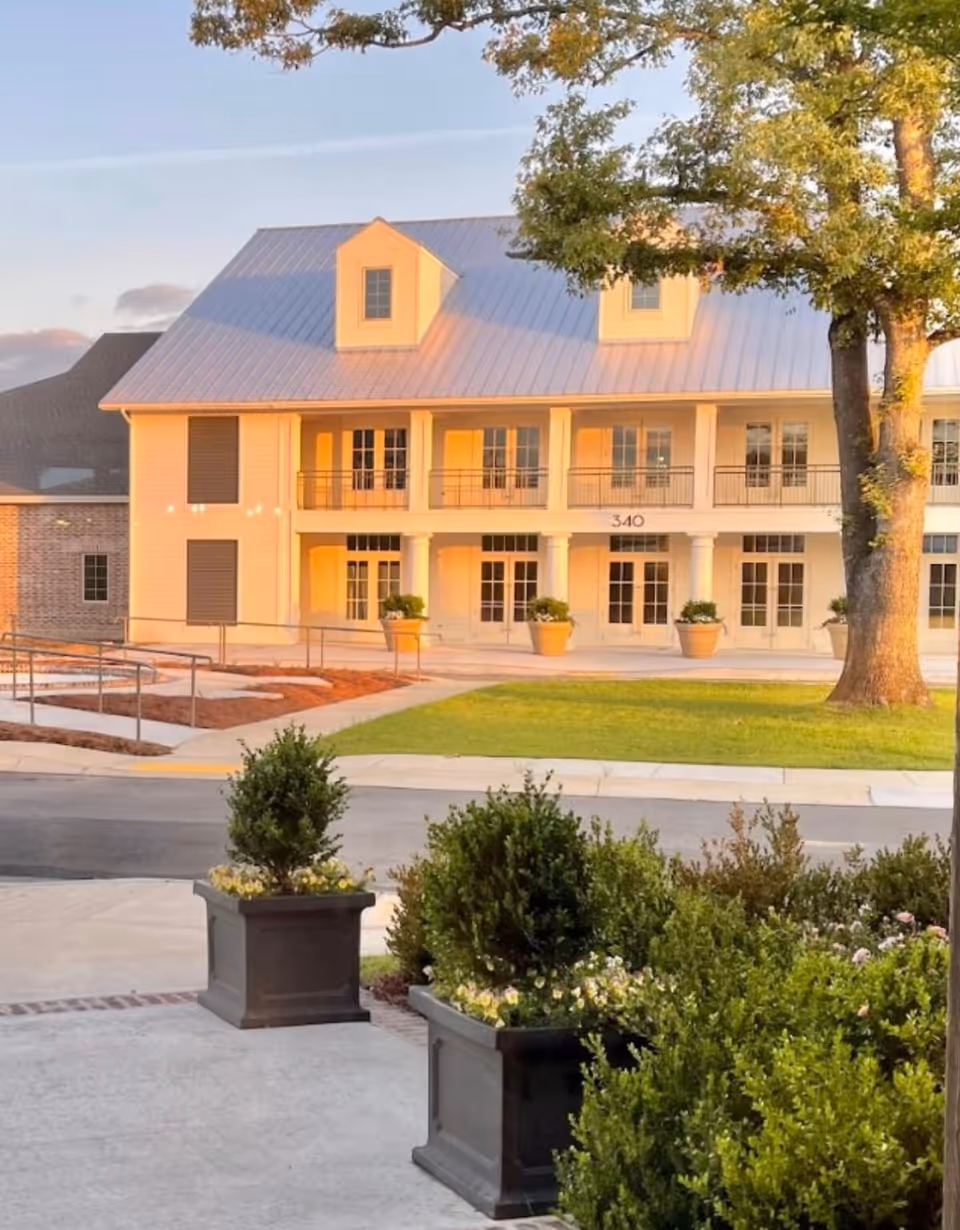 Exterior view of a two-story building with a metal roof and large windows, surrounded by greenery and potted plants, with a tree on the right side and a clear sky above.