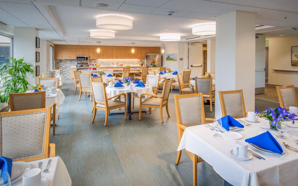Well-lit communal dining room with multiple tables set with white linens and blue napkins, wooden chairs, and a serving counter/kitchenette in the back.