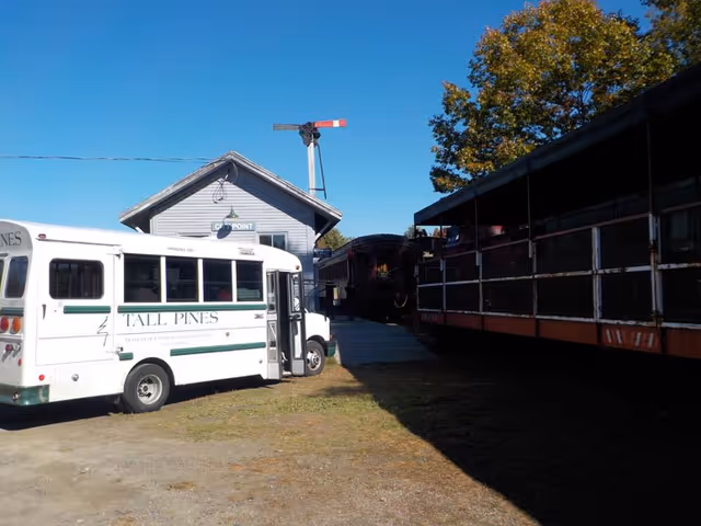 A white Tall Pines Community shuttle bus parked near a small building with a sign that reads 'CUP POINT'. There is a railroad signal above the building and a vintage train car on the right side of the image. The scene is outdoors on a sunny day with a clear blue sky and some trees in the background.
