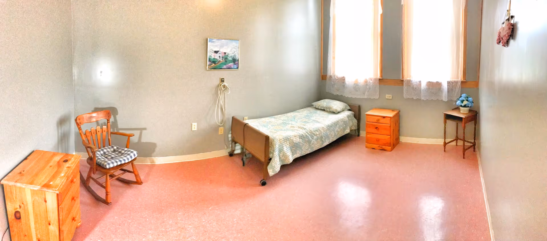 A simple, small bedroom with a single bed covered with a light blue patterned quilt and a pillow. There are two wooden nightstands, one on each side of the bed, and a wooden rocking chair with a checkered cushion. The room has three tall windows with sheer white curtains, a small framed painting on the wall above the bed, and a small table with a blue flower arrangement. The floor is pink linoleum, and the walls are painted light gray.