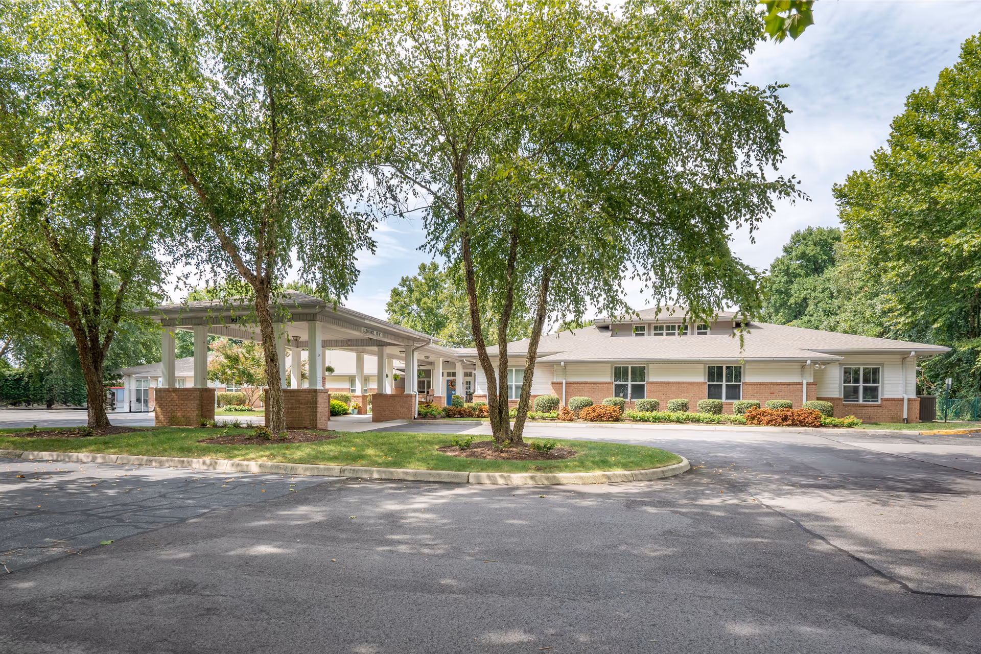 Exterior view of a single-story senior living facility building with a covered entrance, surrounded by trees and landscaping under a partly cloudy sky.