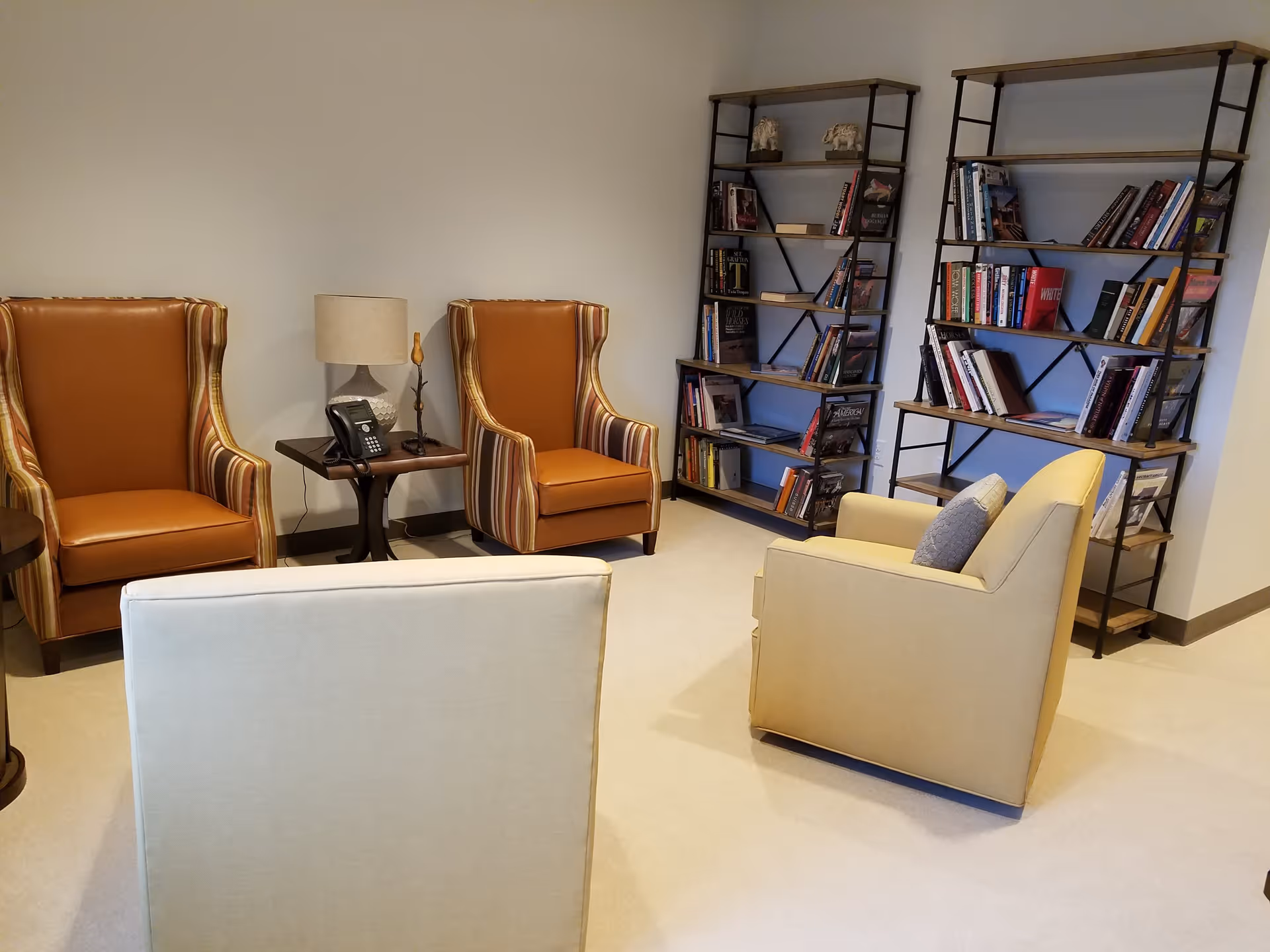 Small sitting area with several armchairs, a side table holding a lamp and phone, and bookshelves filled with books against a wall.