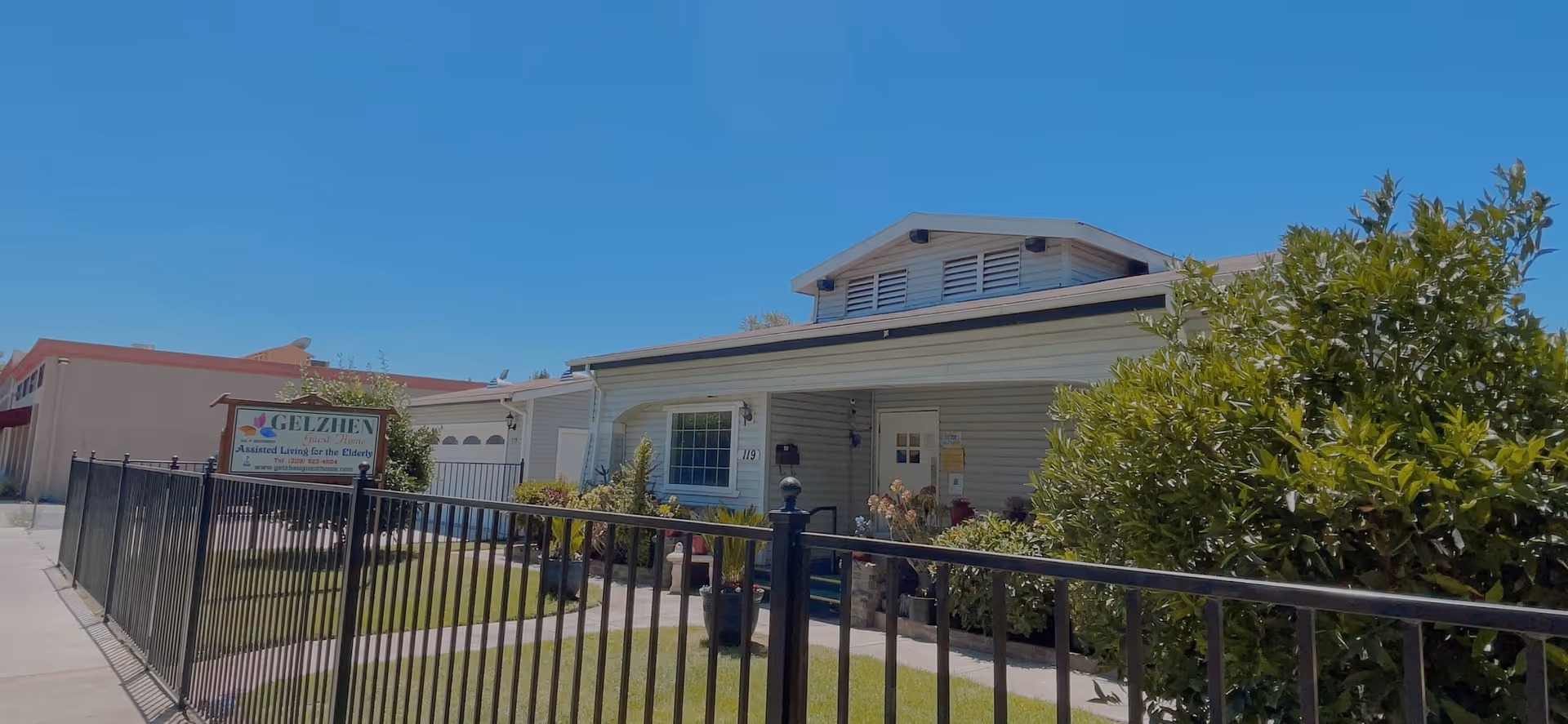Exterior view of Gelzhen Guest Home, a single-story building with light-colored siding and a covered porch. The front yard is enclosed by a black metal fence and features green grass, shrubs, and a sign that reads 'Gelzhen Assisted Living for the Elderly'. The sky is clear and blue.