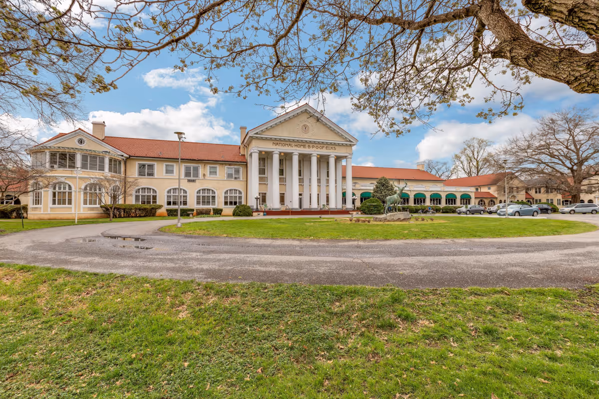 Front facade of a large colonial-style senior living building with tall white columns, a circular driveway, and a lawn with an elk statue.