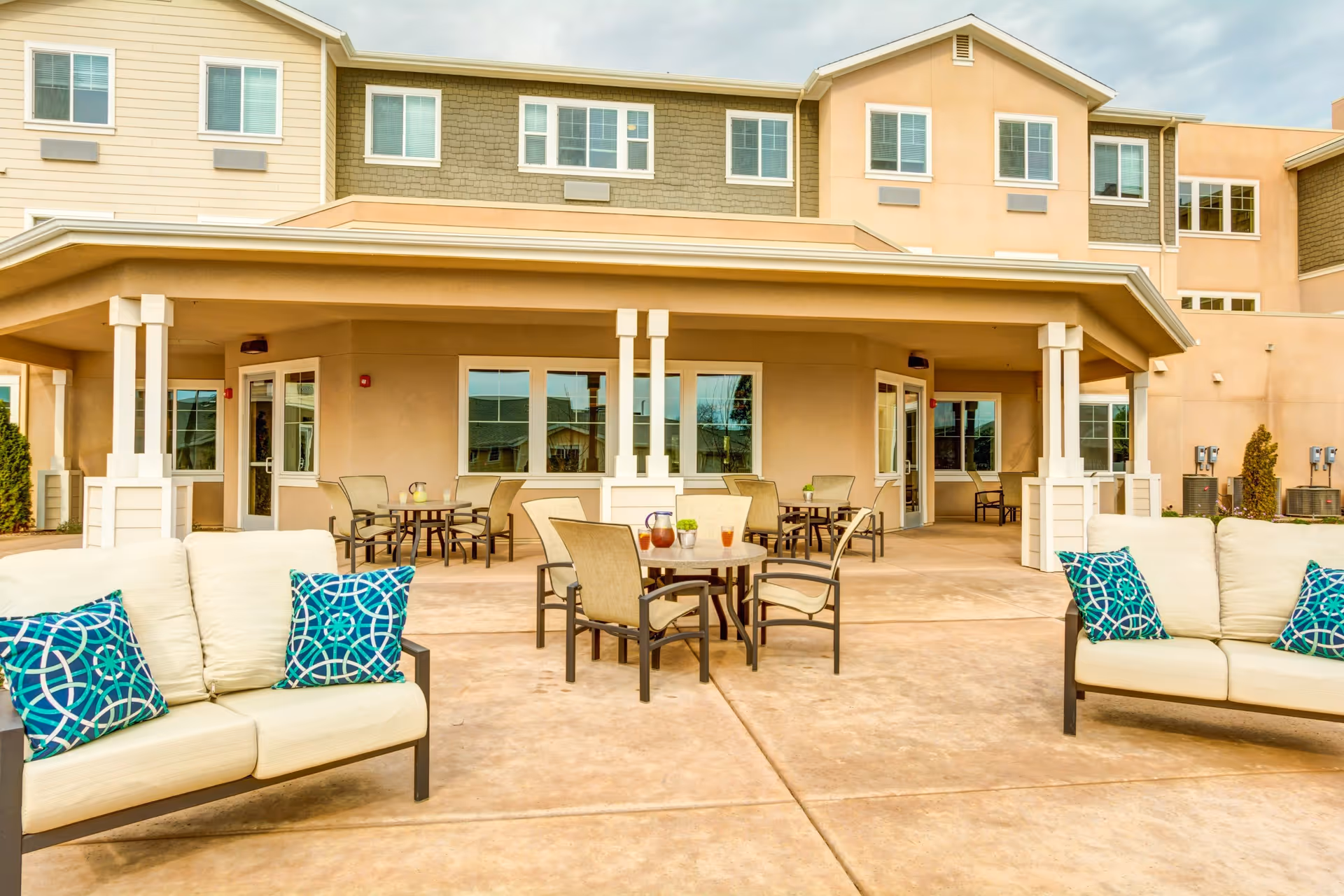 Outdoor patio area of a senior living facility with cushioned seating and tables with chairs. The building has multiple windows and a covered porch area. The patio has beige flooring and blue patterned pillows on the sofas.