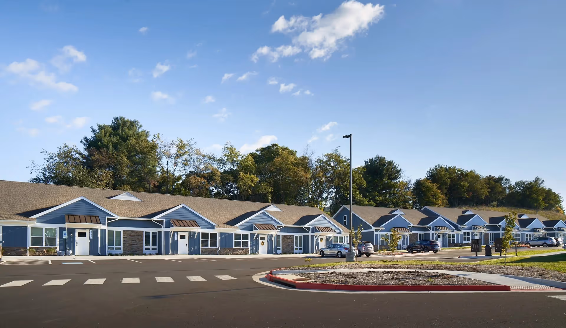 Row of single-story blue residential buildings with parking, a driveway, and trees under a clear blue sky.
