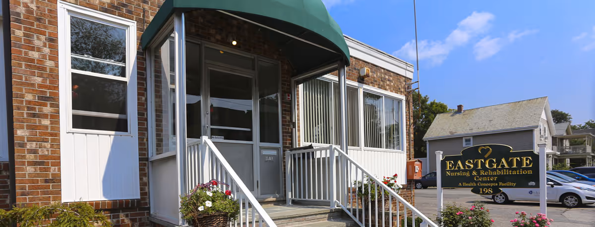 Entrance of Eastgate Nursing & Rehabilitation Center showing a brick building with a green awning over the door, white railings on the steps leading up to the entrance, and a sign with the facility's name and address in front. There are some plants and flowers near the entrance and a parking lot with cars in the background.