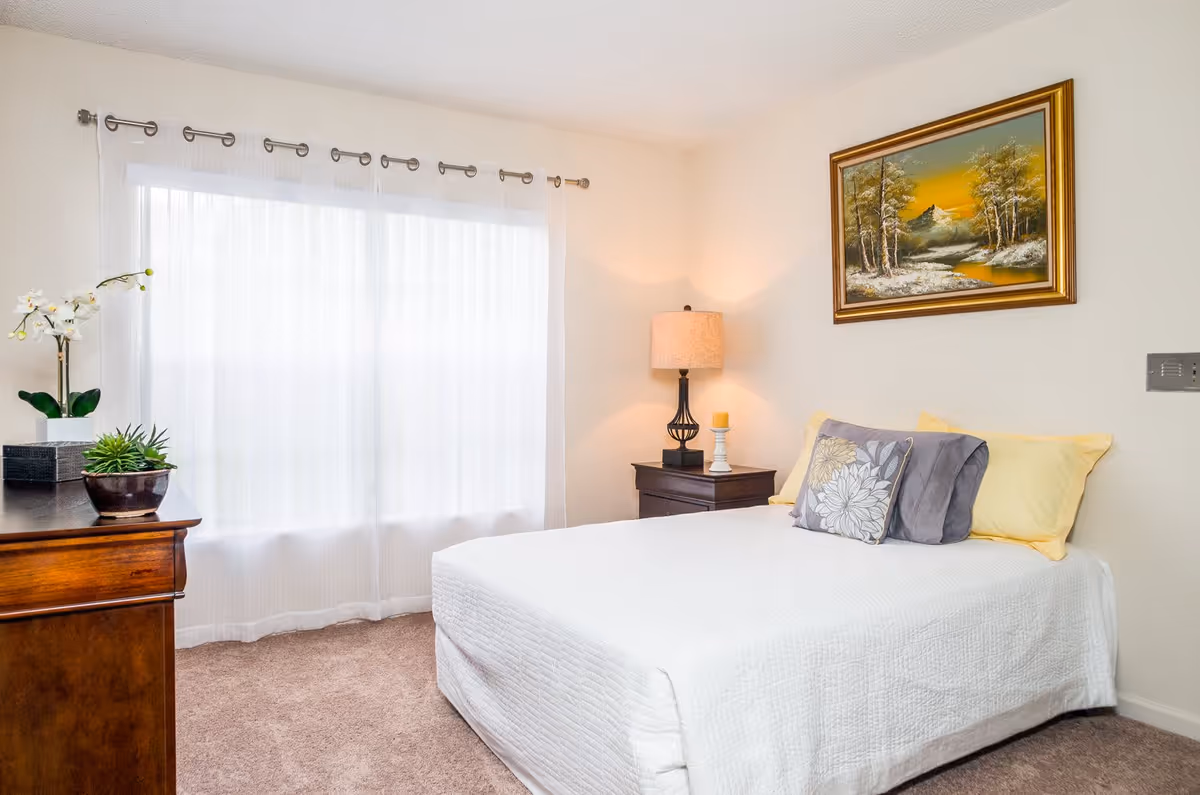 A bright bedroom with a neatly made bed covered in a white quilt and decorated with yellow and gray pillows. To the right of the bed is a dark wooden nightstand with a lamp and a candle. A framed painting of a landscape with trees and a river hangs on the wall above the bed. On the left side of the room, there is a wooden dresser with two potted plants and a sheer white curtain covering a large window.