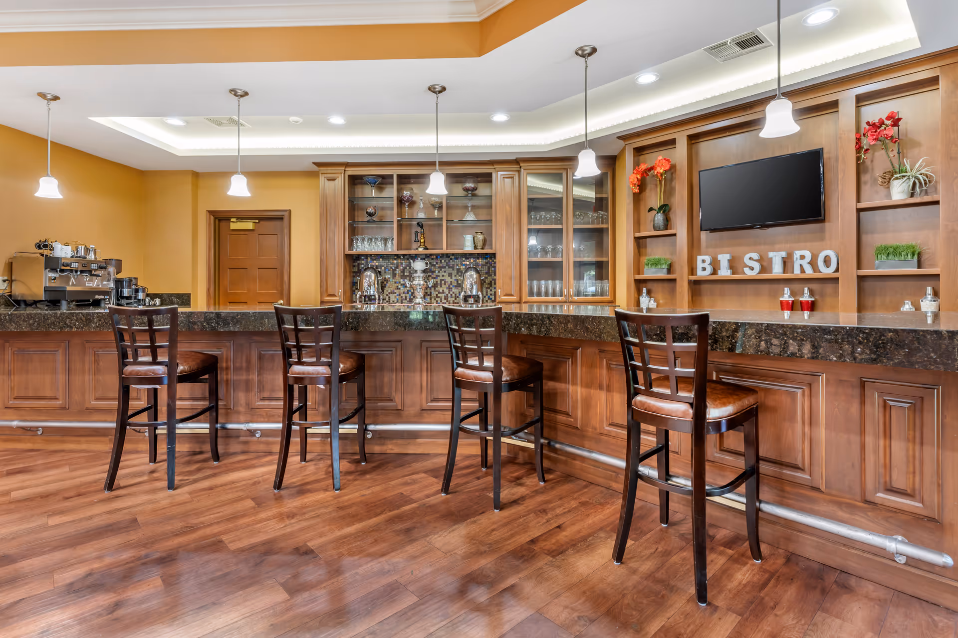 Interior view of a bistro area with a long wooden counter and four high chairs with brown leather seats. Behind the counter are wooden cabinets with glass doors displaying glassware and decorative items. The wall features a mounted flat-screen TV above shelves with plants and decorative letters spelling BISTRO. Pendant lights hang from the ceiling, and the floor is wooden.