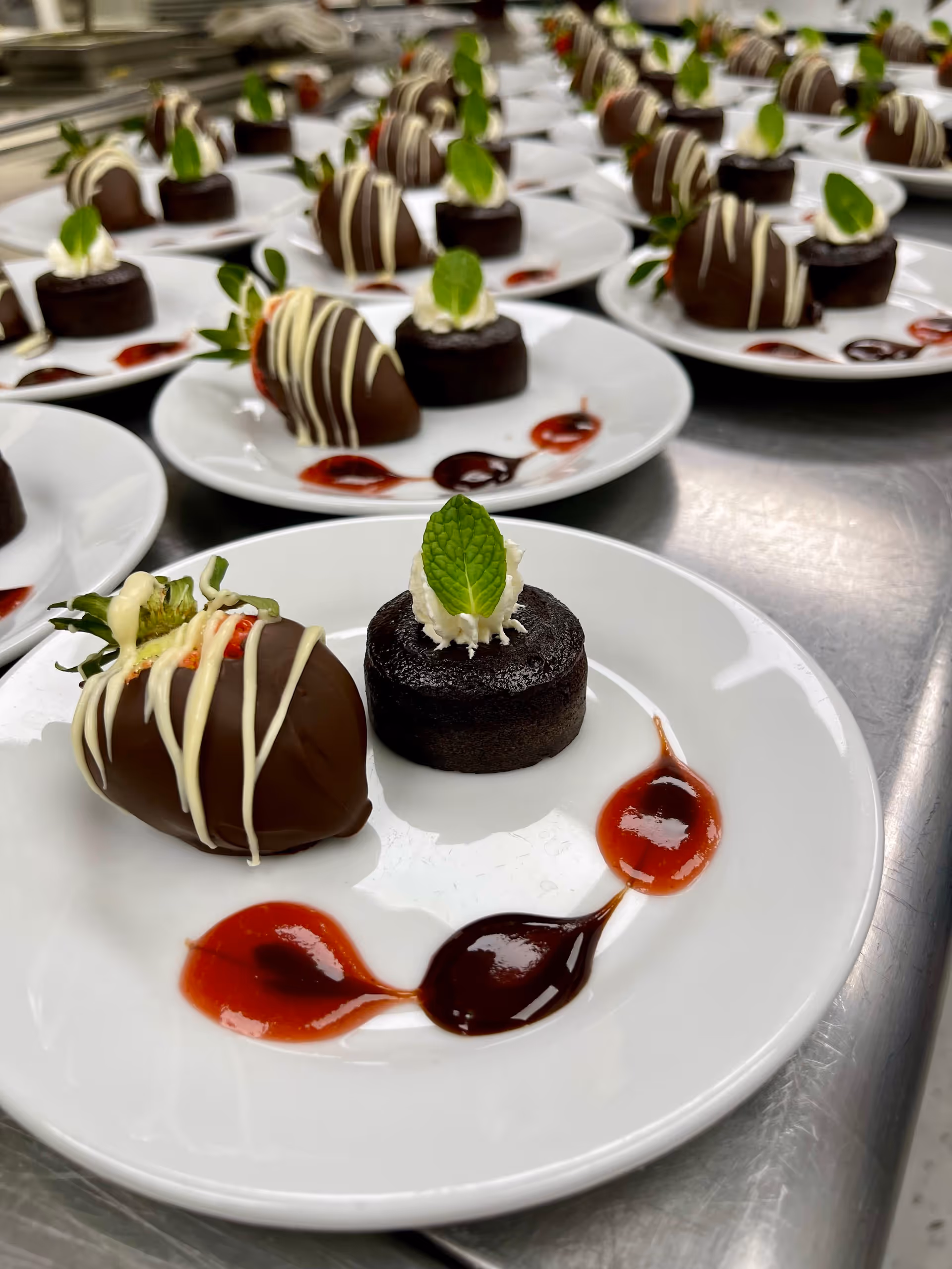 Close-up view of multiple white plates arranged on a metal surface, each plate featuring a chocolate-covered strawberry drizzled with white chocolate, a small chocolate cake topped with whipped cream and a mint leaf, and decorative red and dark sauce dots on the plate.