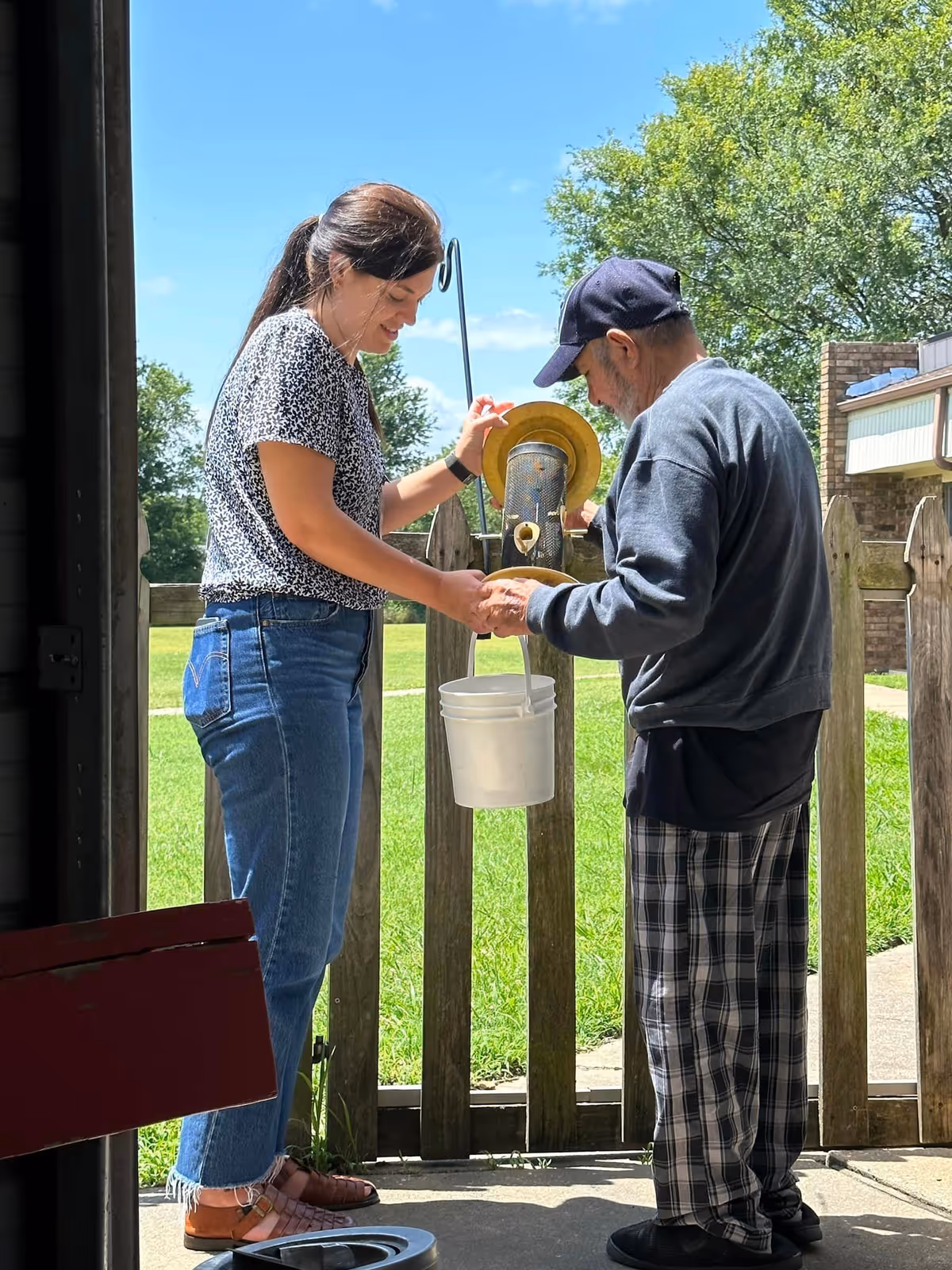 A woman and an elderly man stand outside near a wooden fence on a sunny day. The woman is helping the man fill a white bucket from a yellow bird feeder. The woman is wearing a patterned short-sleeve shirt, blue jeans, and sandals, while the man is dressed in a dark long-sleeve shirt, plaid pants, and a navy blue cap. Green grass and trees are visible in the background.