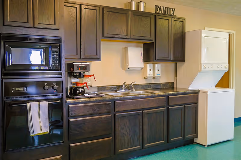 A kitchen area with dark wooden cabinets, a black microwave and oven, a coffee maker with two coffee pots, a double sink, paper towel and soap dispensers mounted on the wall, and a stacked white washer and dryer unit. The word FAMILY is displayed on top of the cabinets.