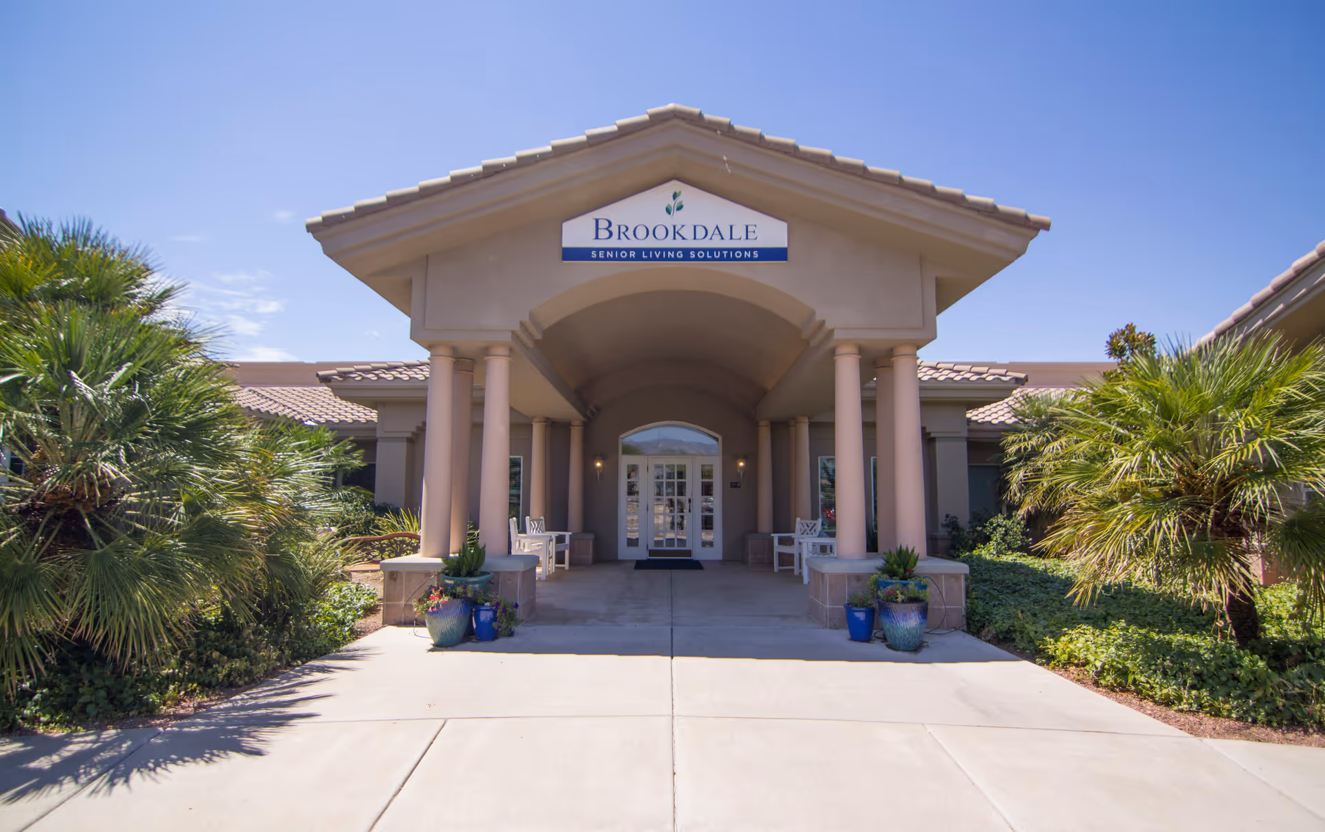 Front entrance of the Brookdale Tanque Verde senior living building with a covered portico, columns, benches, potted plants, and signage.