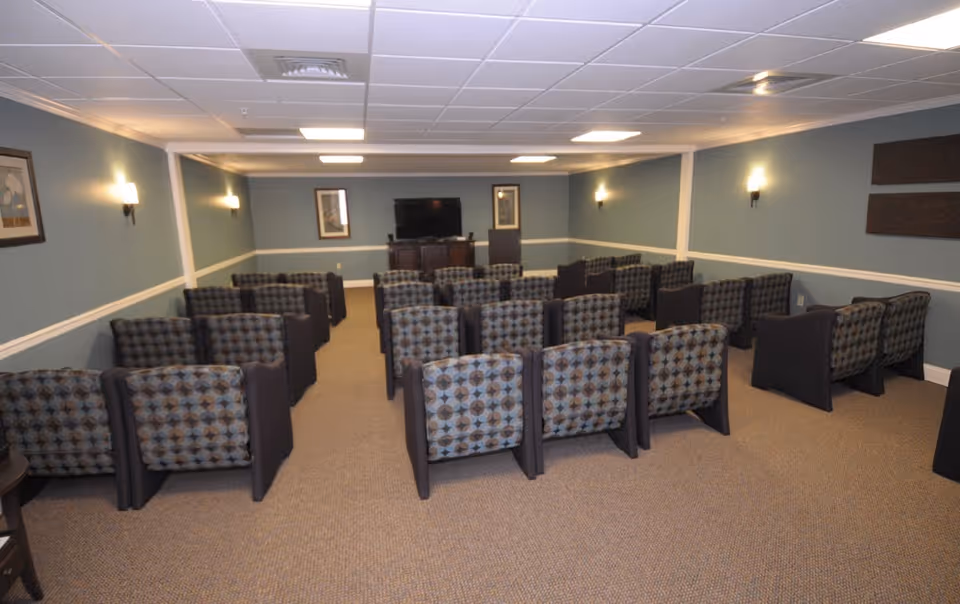 A small community media room with multiple rows of patterned armchairs facing a television at the front.